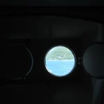 Three round ship portholes in a dark cabin wall, only the center one open to reveal a bright view of the sea and coastline beyond