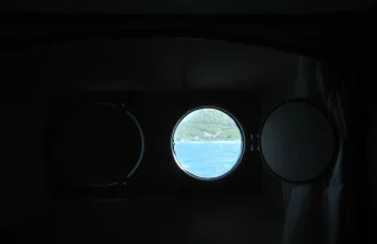 Three round ship portholes in a dark cabin wall, only the center one open to reveal a bright view of the sea and coastline beyond
