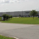 A real Amazon fulfillment center building with the Amazon logo, a chain-link fence, and a blue sign reading 'No personal or unauthorized vehicles beyond this point' in the foreground