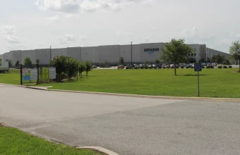A real Amazon fulfillment center building with the Amazon logo, a chain-link fence, and a blue sign reading 'No personal or unauthorized vehicles beyond this point' in the foreground
