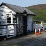 A small white wooden toll booth with a Pay Point sign and a fare board at Penmaenpool Toll Bridge, with orange traffic cones on the bridge deck