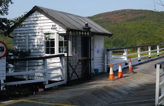 A small white wooden toll booth with a Pay Point sign and a fare board at Penmaenpool Toll Bridge, with orange traffic cones on the bridge deck