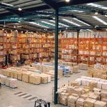 Aerial view inside a large warehouse with tall orange pallet racking fully stocked with boxed goods, workers and forklifts moving among the aisles