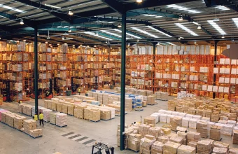 Aerial view inside a large warehouse with tall orange pallet racking fully stocked with boxed goods, workers and forklifts moving among the aisles