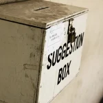 A weathered wooden suggestion box labeled SUGGESTION BOX, mounted on a wall with a padlock and a slot for messages