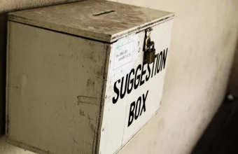 A weathered wooden suggestion box labeled SUGGESTION BOX, mounted on a wall with a padlock and a slot for messages