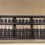 A dark wooden abacus with two rows of black beads on copper-colored rods, resting on a clear acrylic stand against a beige wall