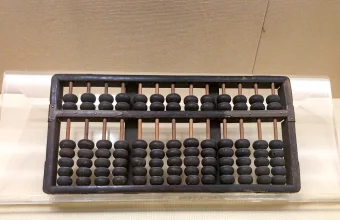 A dark wooden abacus with two rows of black beads on copper-colored rods, resting on a clear acrylic stand against a beige wall