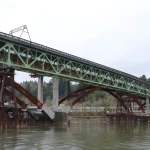 The Angular 2 Rewrite Turns a Migration Lesson Into a Security Bill: AngularJS Still Draws 533,480 Downloads a Week, and 10 Advisories Have No Open-Source Fix An old green steel truss bridge resting on temporary supports beside a new arch bridge under completion on a river in Portland, Oregon