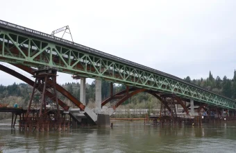 An old green steel truss bridge resting on temporary supports beside a new arch bridge under completion on a river in Portland, Oregon