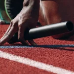 A relay baton lying on a red running track beside a sprinter’s hand at the starting blocks