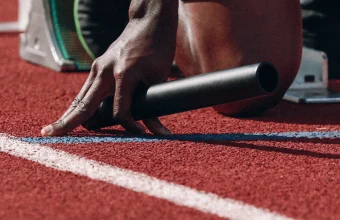 A relay baton lying on a red running track beside a sprinter’s hand at the starting blocks