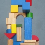 Colorful wooden toy blocks stacked into a tower with an arch, a tall blue pillar and a red triangle against a gray-blue wall