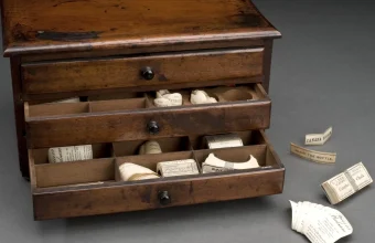 A small wooden label cabinet with three drawers, the lower two pulled open to show compartments of paper labels, with loose labels scattered on a grey surface beside it
