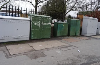 A row of green and grey fibre broadband street cabinets on a pavement beside a fence in Iver, England
