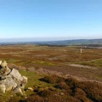 A pile of grey stones forming a cairn on open heather moorland under a clear blue sky