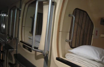 Row of capsule hotel pods with white pillows and folded blankets, each capsule an idle sleeper packed into a shared rack