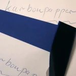 A handwritten word on white paper with a sheet of blue carbon paper peeled back to show a faint carbon copy of the same word underneath