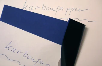 A handwritten word on white paper with a sheet of blue carbon paper peeled back to show a faint carbon copy of the same word underneath