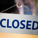 A white CLOSED sign with blue letters hanging from a suction cup and chain on a glass shop door
