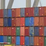 A wall of stacked, multicolored shipping containers at the Port of Rotterdam with port crane legs rising behind them