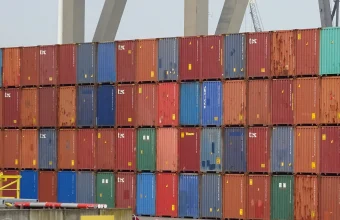 A wall of stacked, multicolored shipping containers at the Port of Rotterdam with port crane legs rising behind them
