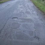 A rural road with many patched asphalt repairs and one open pothole