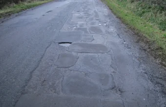 A rural road with many patched asphalt repairs and one open pothole
