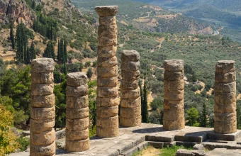 Six weathered stone columns of the Temple of Apollo at Delphi, Greece, standing on a stone platform above a valley of olive groves