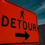 An orange DETOUR road sign with a pedestrian symbol and a right-pointing arrow against a dark blue sky