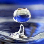 A single drop of water hovering above a rippling blue water surface, with a small column of water rising beneath it