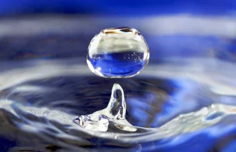 A single drop of water hovering above a rippling blue water surface, with a small column of water rising beneath it