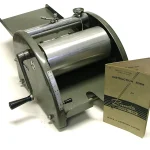 A vintage Banda Model Ten spirit duplicator, a hand-cranked paper copying machine, shown with its instruction book on a white background