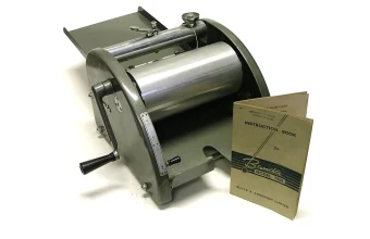 A vintage Banda Model Ten spirit duplicator, a hand-cranked paper copying machine, shown with its instruction book on a white background