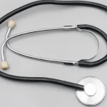 A black-tubed stethoscope with cream ear tips and a steel chest piece laid flat on a light gray surface