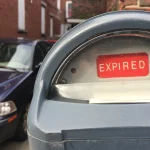 A gray parking meter with a red EXPIRED flag in its window and a dark parked car blurred behind it