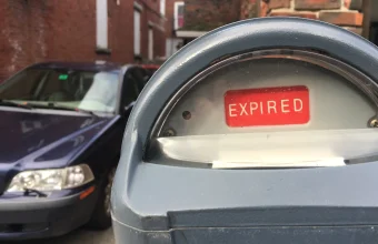 A gray parking meter with a red EXPIRED flag in its window and a dark parked car blurred behind it