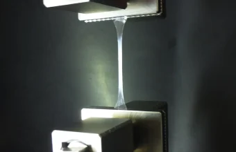 A thin transparent polymer test specimen stretched between the metal grips of a tensile testing machine