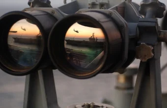 Large naval binoculars whose two lenses each reflect the same sunset scene of a ship and a helicopter