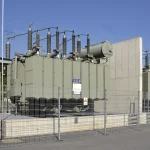 A tall concrete fire wall between two large transformers behind a metal fence at the Kachlet power station in Passau, Germany