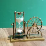 A bronze-framed hourglass with white sand beside a small Ferris wheel ornament on a wooden table against a teal wall