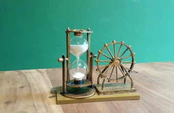 A bronze-framed hourglass with white sand beside a small Ferris wheel ornament on a wooden table against a teal wall