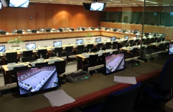 An empty interpreters booth with a microphone console and monitors overlooking a large Council meeting room in Brussels