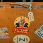 A worn leather suitcase with a paper hang tag and several vintage hotel luggage labels stuck to it