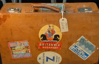 A worn leather suitcase with a paper hang tag and several vintage hotel luggage labels stuck to it