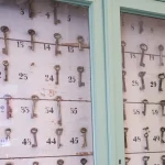 A glass-fronted green cabinet holding rows of old keys on numbered hooks, with several hooks empty
