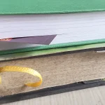 Two stacked hardcover books on a table, the lower one with a yellow ribbon bookmark hanging from its pages and the upper one with a paper bookmark sticking out