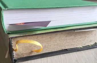 Two stacked hardcover books on a table, the lower one with a yellow ribbon bookmark hanging from its pages and the upper one with a paper bookmark sticking out
