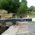 A black and white wooden lock-gate balance beam beside a canal lock chamber on the Stratford-upon-Avon Canal, with a sign reading Keep boat forward of cill marker