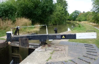 A black and white wooden lock-gate balance beam beside a canal lock chamber on the Stratford-upon-Avon Canal, with a sign reading Keep boat forward of cill marker